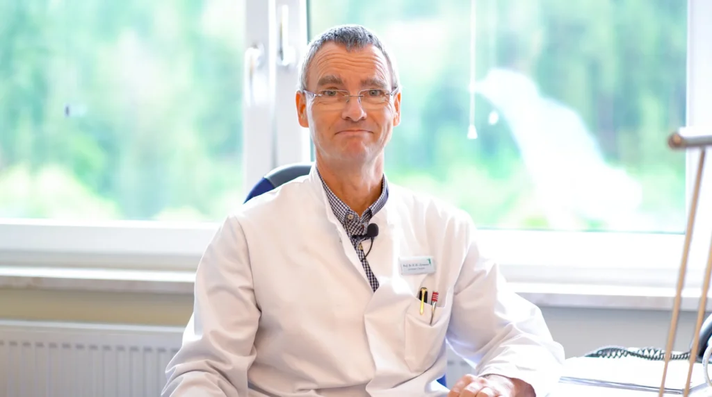 Die Hauptperson des Bildes ist ein Mann mittleren Alters, der eine Brille und einen weißen Laborkittel trägt. Die Person ist vor einem Fenster positioniert, das einen verschwommenen Blick auf grüne Natur bietet. Die Person trägt ein Gitterhemd unter dem Laborkittel. Ein Namensschild sticht hervor, das "Professor Dr. D-H. Zermann" anzeigt, gefolgt unter seinem Namen von "Leitender Chefarzt". In der Brusttasche befinden sich zwei Stifte. Einer davon ist golden und der andere hat gold und rot als Farben. Die Person sitzt an einem Schreibtisch, auf dem ein Telefon und ein dreibeiniges Gerät zu sehen sind. Für das Telefon ist nur die Basis und das Kabel sichtbar. An seinem Hemd ist ein Mikrofon angebracht, was darauf hindeutet, dass er spricht, aber nicht zu sehen ist, was er sagt. Die Gesamtstimmung ist professionell und akademisch.