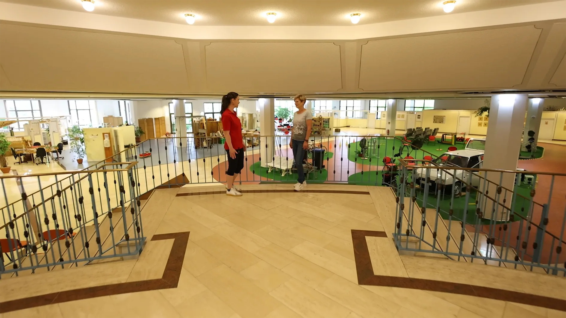 Das Bild zeigt eine Innenansicht eines großen, weitläufigen Raumes, der offenbar als Ausstellungs- oder Lernbereich dient. Im Vordergrund stehen oben auf einer Balustrade, die einen Blick nach unten auf den Hauptbereich ermöglicht, zwei Frauen. Eine Frau trägt ein rotes Shirt und schwarze Hosen, die andere ein graues T-Shirt und Jeans. Der Hauptbereich unten ist in verschiedene Zonen unterteilt, die unterschiedliche Funktionen oder Exponate zeigen.