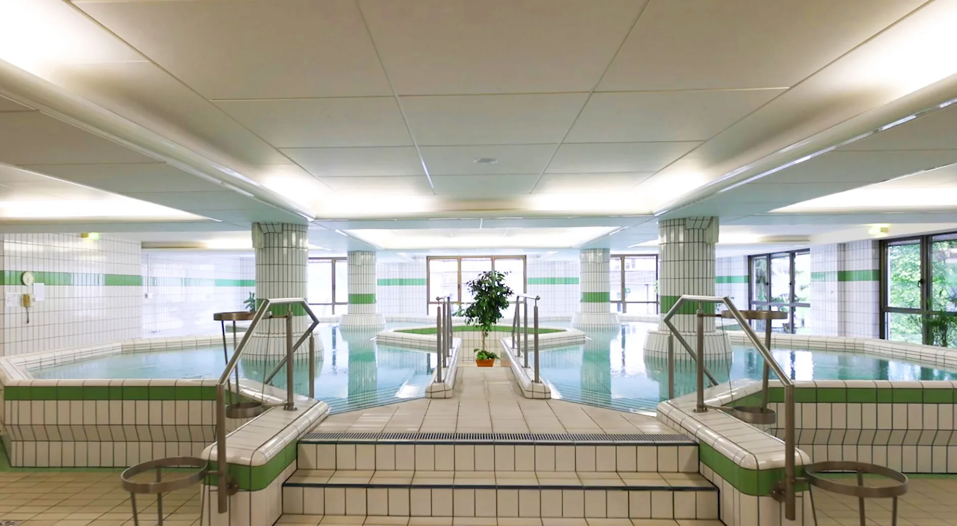 Das Bild zeigt eine Innenansicht eines großen Schwimmbads oder Kurzentrum-Bereichs. Im Vordergrund befinden sich Stufen, die zu einem gefliesten Bereich führen, umgeben von Schwimmbecken mit klarem Wasser. Die Wände der Becken und der Umgebung sind großflächig mit weißen Kacheln versehen, unterbrochen von grünen Streifen. Vier große, runde Säulen mit ähnlicher grüner und weißer Fliesenverkleidung stützen die weiße Decke, die mit langen Leuchtstoffröhren gut beleuchtet ist. In der Mitte der Anlage befindet sich eine kleine Insel mit einer Topfpflanze. Im Hintergrund sind große Fenster zu sehen, die Tageslicht hereinlassen und einen Blick nach draußen auf Bäume und Gebäude ermöglichen. Mehrere Edelstahlhandläufe und kleine Tische verteilen sich im Raum. Die Farbgebung ist hauptsächlich weiß und grün.