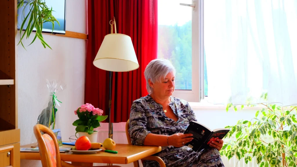 Eine Frau mit kurzen, grauen Haaren sitzt in einem Wohnraum und liest ein Buch. Sie trägt ein gemustertes Kleid. In der Szene sind mehrere Pflanzen, ein Tisch mit Obst und eine Stehlampe zu sehen. Durch ein Fenster im Hintergrund blickt man auf eine grüne Landschaft.