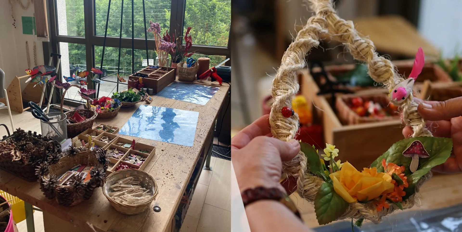 Das Bild zeigt einen Bastel- oder Werkraum mit einem großen Holztisch im Vordergrund, der mit vielfältigen Handwerksmaterialien und Utensilien übersät ist. Dazu gehören Körbe mit Tannenzapfen, Schachteln mit kleinen Holzobjekten, Dekomaterialien, eine Heißklebepistole, Scheren und verschiedene Bastelarbeiten. Im Hintergrund ist ein großes Fenster zu sehen, das eine Aussicht auf einen dichten Wald bietet. Ein Garderobenständer und diverse weitere kleinere Gegenstände sind ebenfalls erkennbar. An einer Wand hängen Makramee-Arbeiten und Notizen.