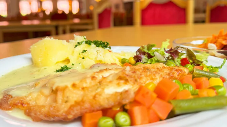 Das Bild zeigt eine Nahaufnahme eines Gerichts auf einem weißen Teller, bestehend aus einem paniertem Stück Fleisch oder Fisch, das mit einer hellen Soße übergossen ist, dazu Kartoffelpüree, gemischtes Gemüse (Erbsen, Karotten, grüne Bohnen, Mais) und ein kleiner Salat. Der Hintergrund ist unscharf und zeigt einen Innenbereich, möglicherweise ein Restaurant oder eine Kantine, mit Tischen und roten Stühlen.