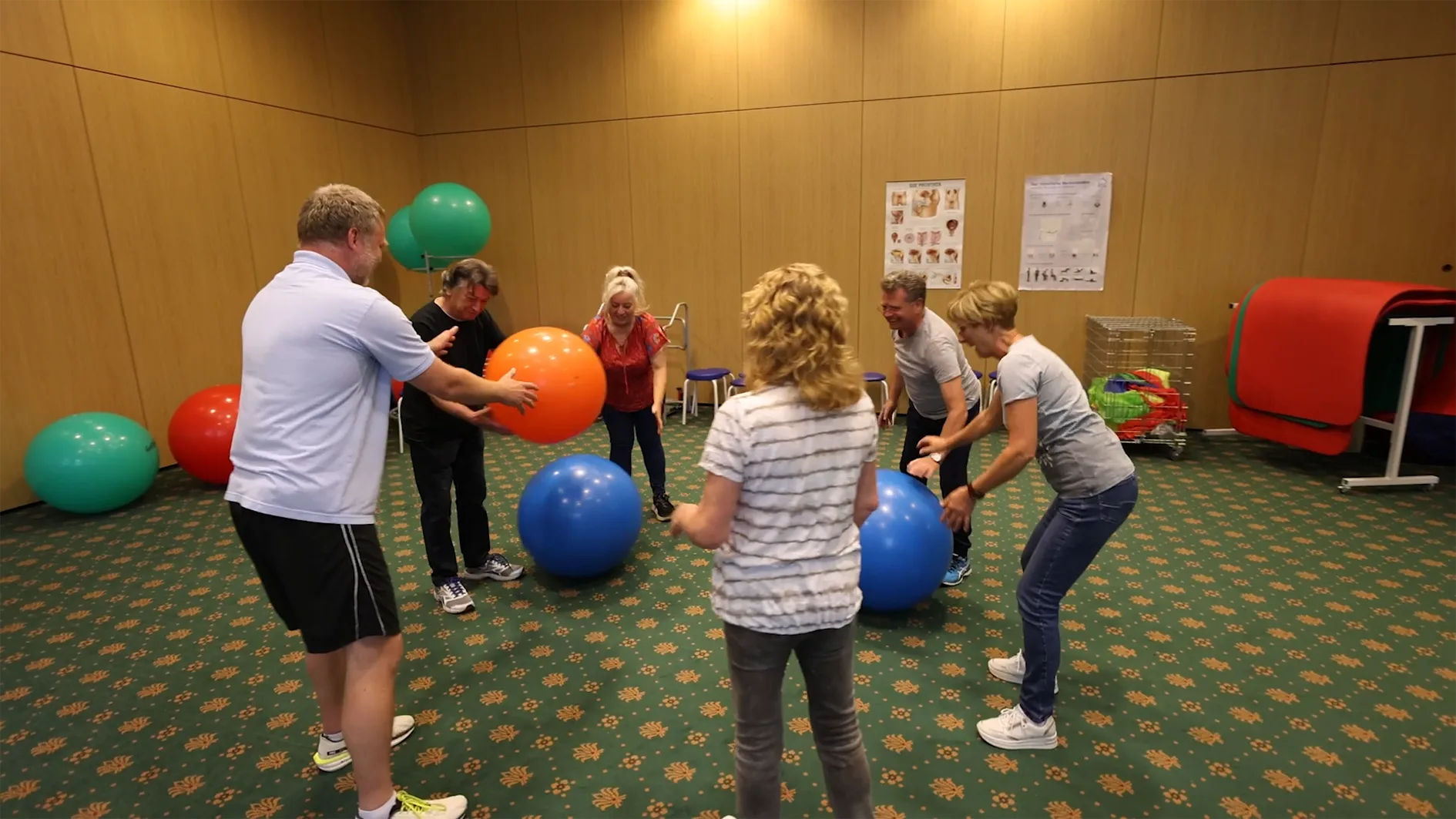 Eine Gruppe von sieben Personen in einem großen Raum mit Holzwänden und grünem Teppichboden nimmt an einer Übungseinheit mit Gymnastikbällen teil. Während einige Bälle auf dem Boden liegen, werden andere gerade von den Teilnehmern bewegt. Im Hintergrund sind auf Plakaten schematische Darstellungen des menschlichen Körpers zu sehen.