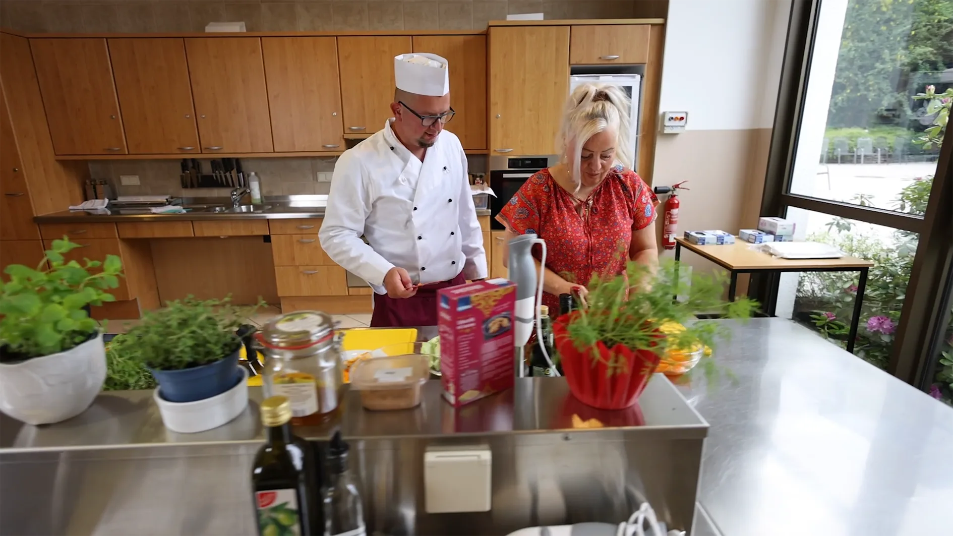 Dieses Bild zeigt einen Koch und eine Frau in einer hell beleuchteten Küche mit Holzschränken und großen Fenstern. Sie scheinen beim Kochen oder Zubereiten von Speisen zu sein, da Utensilien und Zutaten auf den Edelstahlarbeitsflächen zu sehen sind. Pflanzen, wie Kräuter, sind ebenfalls auf dem Tisch platziert.