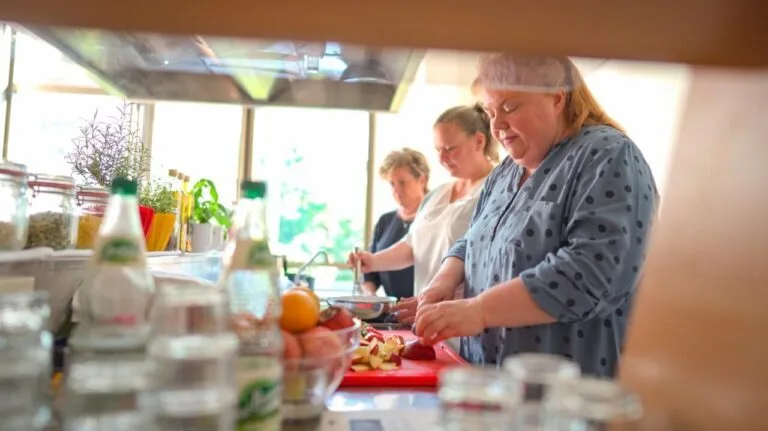 Das Bild zeigt mehrere Frauen, die in einer Küche gemeinsam Speisen zubereiten. Sie sind mit verschiedenen Aufgaben beschäftigt, wie dem Kleinschneiden von Obst und dem Rühren in Töpfen. Im Vordergrund sind Flaschen und Gläser zu sehen, die auf einer Arbeitsfläche stehen.