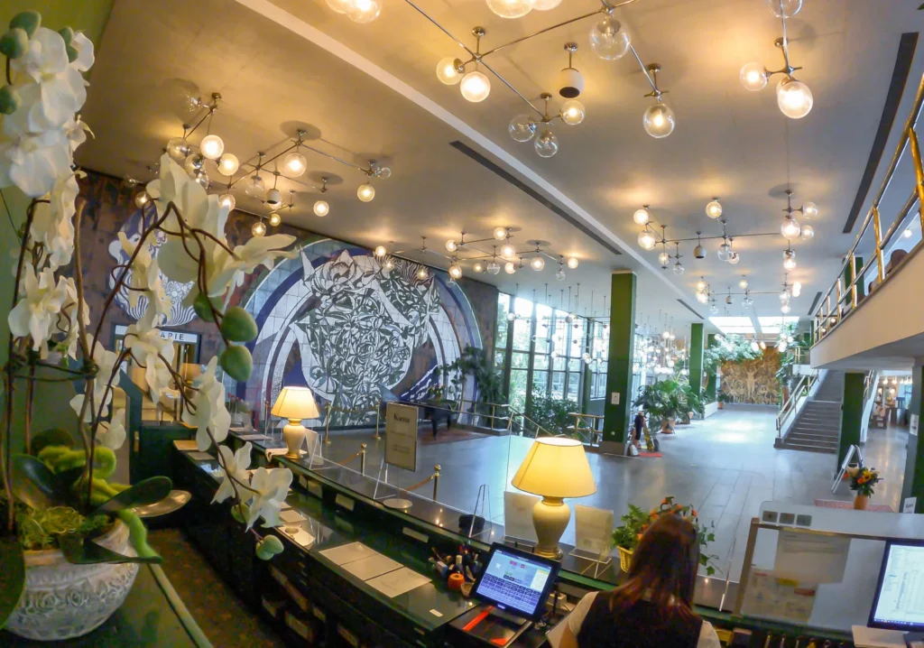 Das Bild zeigt die Empfangshalle eines Gebäudes, möglicherweise eines Hotels oder einer Kultureinrichtung. Im Vordergrund, links, ist eine große Pflanze mit weißen Blüten zu sehen, die zum Design der Halle beiträgt. Rechts befindet sich ein Empfangstresen mit einer Person dahinter und einem Tablet-Computer auf dem Tresen. Eine Tischlampe mit einem hellen Schirm steht ebenfalls auf dem Tresen. Im Hintergrund dominiert eine große Wandmalerei mit einem abstrakten, geometrischen Muster in Blautönen und Weiß. Die Decke ist mit zahlreichen kugelförmigen Lampen in unterschiedlichen Größen ausgestattet, die ein warmes Licht abgeben. Auf der rechten Seite des Bildes ist eine Treppe sichtbar, die in ein Obergeschoss führt, und in der Ferne sind auch große Fenster zu erkennen, die Tageslicht hereinlassen. Überall in der Halle stehen Grünpflanzen in Töpfen, die dem Raum eine lebendige Atmosphäre verleihen.