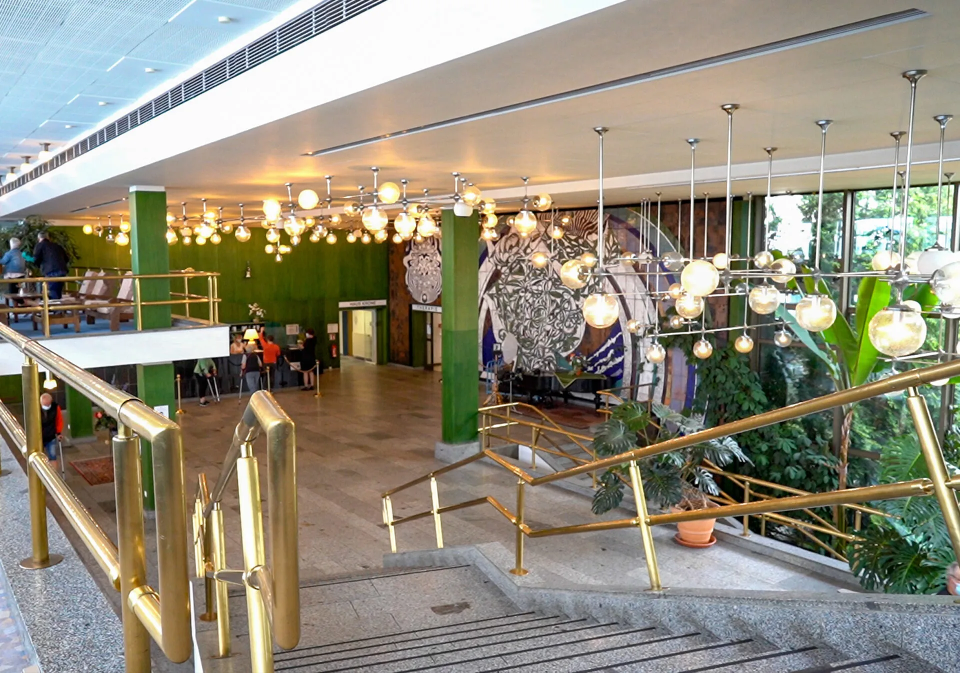 Das Bild zeigt die Innenansicht einer großen Halle oder Empfangsbereich. Im Vordergrund befindet sich eine Treppe mit goldenen Geländern, die nach unten führt. Links davon erstreckt sich ein langer Gang mit gemustertem Teppich. Im oberen Bereich des Bildes ist eine erhöhte Plattform mit Sitzgelegenheiten zu sehen, auf der sich einige Personen aufhalten. Der Hintergrund der Halle ist geprägt von großen, grünen Wänden und beeindruckenden Wandgemälden mit floralen und ornamentalen Mustern. Eine Vielzahl von hängenden Kugelleuchten erhellt den Raum und erzeugt eine warme Atmosphäre. Große Fenster auf der rechten Seite lassen Tageslicht herein und geben den Blick auf üppige Pflanzen im Freien frei. Mehrere Menschen sind in der gesamten Halle verteilt und scheinen sich zu bewegen oder aufzuhalten.