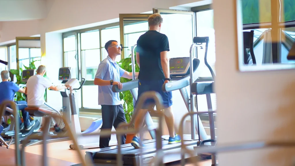 Das Bild zeigt zwei Männer im Fitnessstudio. Einer der Männer ist der Trainer und der andere ist der Klient, der auf einem Laufband trainiert. Zwei weitere Männer sind im Hintergrund zu sehen, die auf Fahrradergometern sitzen. Das Fitnessstudio verfügt über große Fenster, durch die Tageslicht einfällt.