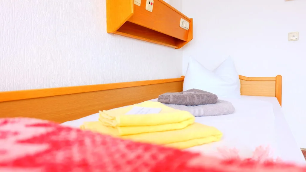 Das Bild zeigt ein sauberes, einfach eingerichtetes Schlafzimmer oder Hotelzimmer. Im Vordergrund ist ein Bett mit weißer Bettwäsche zu sehen, auf dem ordentlich gefaltete gelbe und graue Handtücher liegen. Im Hintergrund befindet sich eine weiße Wand mit einer hölzernen Kopfleiste und einem darüber angebrachten Regal oder einer Leiste mit Steckdosen und Schaltern.