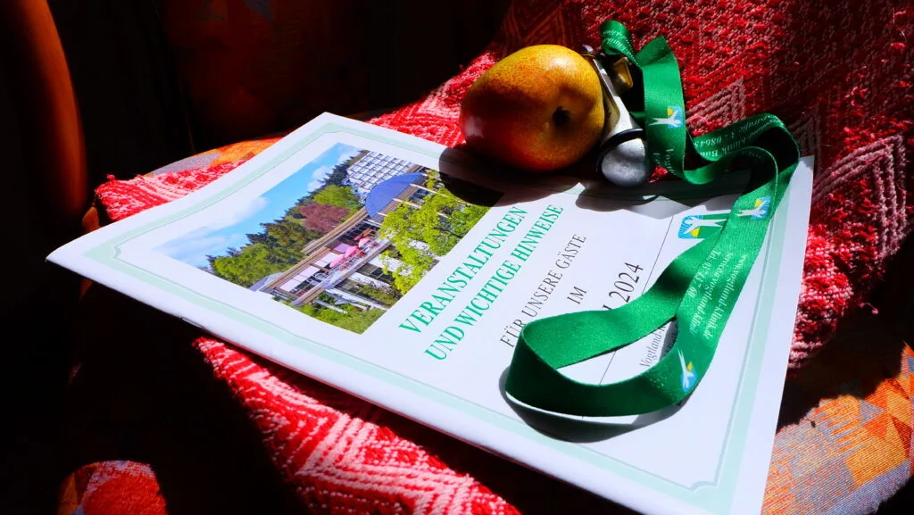 Das Bild zeigt ein Informationsheft mit dem Titel "VERANSTALTUNGEN UND WICHTIGE HINWEISE FÜR UNSERE GÄSTE IM [Monat] 2024", das auf einem roten, gestrickten Stoff liegt. Darauf befinden sich eine Birne, ein grüner Schlüsselanhänger mit der Aufschrift "vogtland-klinik", und ein Anhänger in Form einer Metallflöte. Das Heft hat ein Bild einer Gebäudefassade zwischen Bäumen auf der Vorderseite.