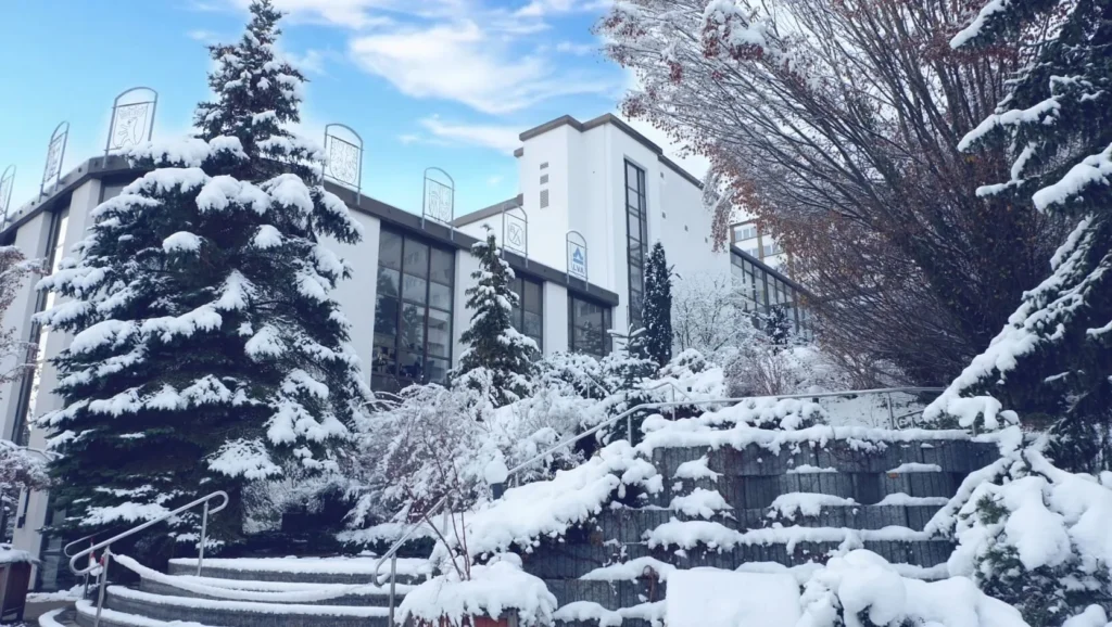 Das Bild zeigt ein weißes Gebäude mit großen Fenstern, umgeben von schneebedeckten Bäumen und Sträuchern. Eine breite, mit Schnee bedeckte Treppe führt zum Gebäude. Der Himmel ist blau mit einigen Wolken. Mehrere Schilder sind auf dem Gebäude zu sehen.