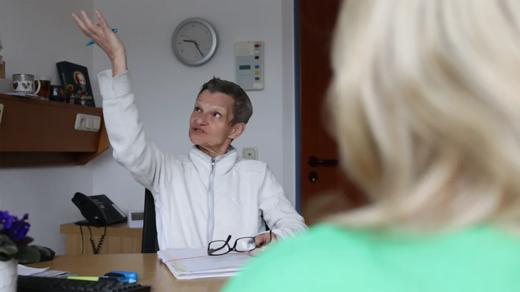 Das Bild zeigt eine Situation in Innenräumen, in der eine Person mit kurzen, grauen Haaren und einem weißen Fleece-Oberteil an einem Schreibtisch sitzt und sich angeregt mit einer anderen Person unterhält, deren blonde Haare und grüne Kleidung im