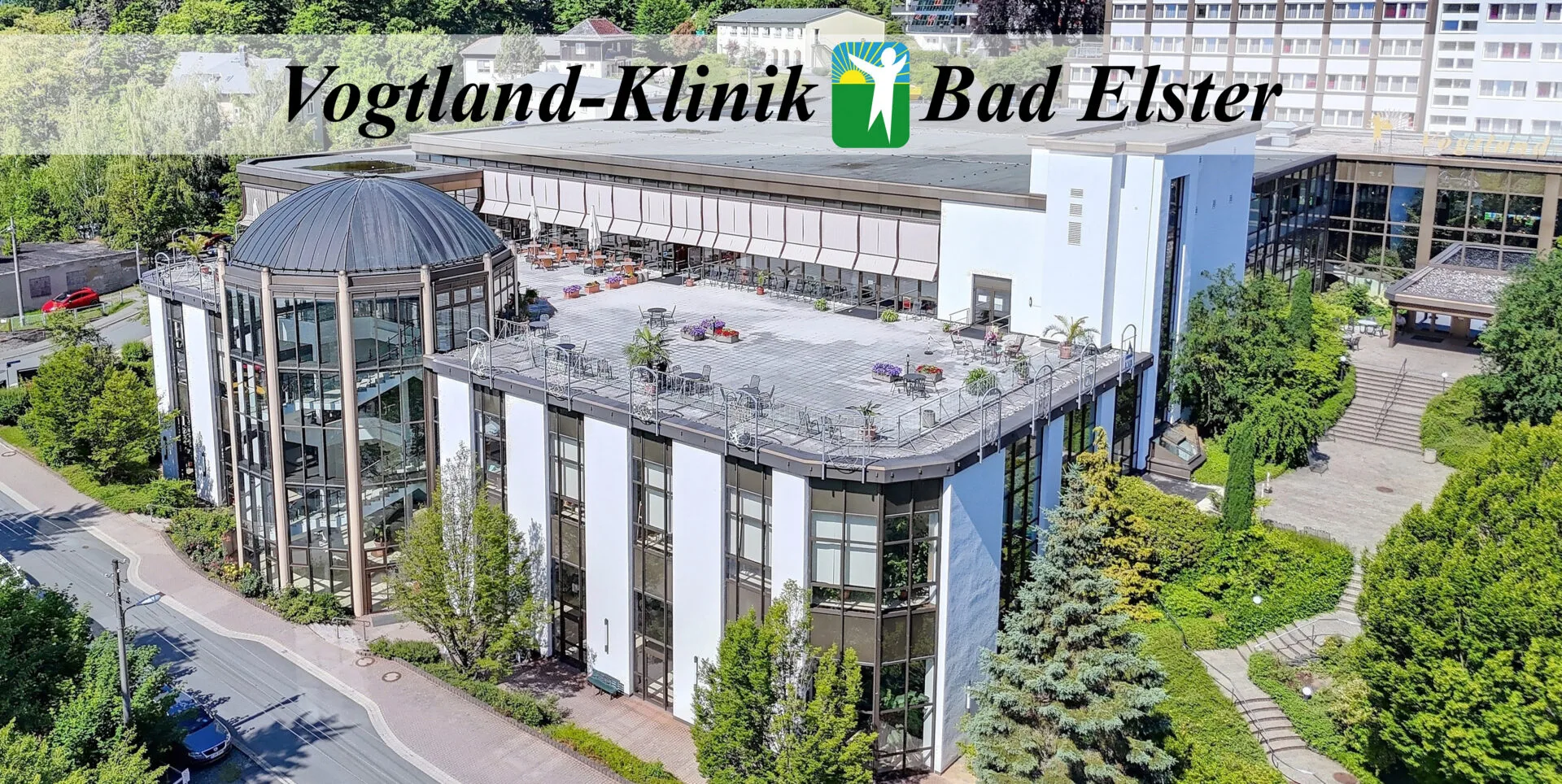 Luftaufnahme der Vogtland-Klinik Bad Elster, ein modernes Gebäude mit Glasfassaden, einer Kuppel und einer Dachterrasse. Die Klinik ist von Bäumen und Grünflächen umgeben, mit Wegen und Treppen. Das Kliniklogo und der Name sind im Bild zu sehen.
