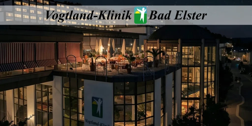 Dieses Bild zeigt die Vogtland-Klinik in Bad Elster bei Nacht. Das Gebäude ist modern und architektonisch ansprechend gestaltet, mit großen Glasfassaden, die hell erleuchtet sind. Besonders auffällig ist die großzügige Dachterrasse im ersten Stock, die mit Tischen, Stühlen und Sonnenschirmen ausgestattet ist und eine einladende Atmosphäre schafft. Im Hintergrund ist ein größeres, mehrstöckiges Gebäude zu sehen, das ebenfalls beleuchtet ist und den Hintergrund der Klinik bildet. Das Logo der Klinik, eine stilisierte menschliche Figur, ist sowohl auf dem Gebäude als auch in einem Banner am oberen Bildrand prominent platziert. Die Szene vermittelt eine ruhige, professionelle und zugleich gastfreundliche Stimmung. Die nächtliche Beleuchtung betont die klaren Linien der Architektur und lässt das Gebäude insgesamt sehr gepflegt und modern wirken.
